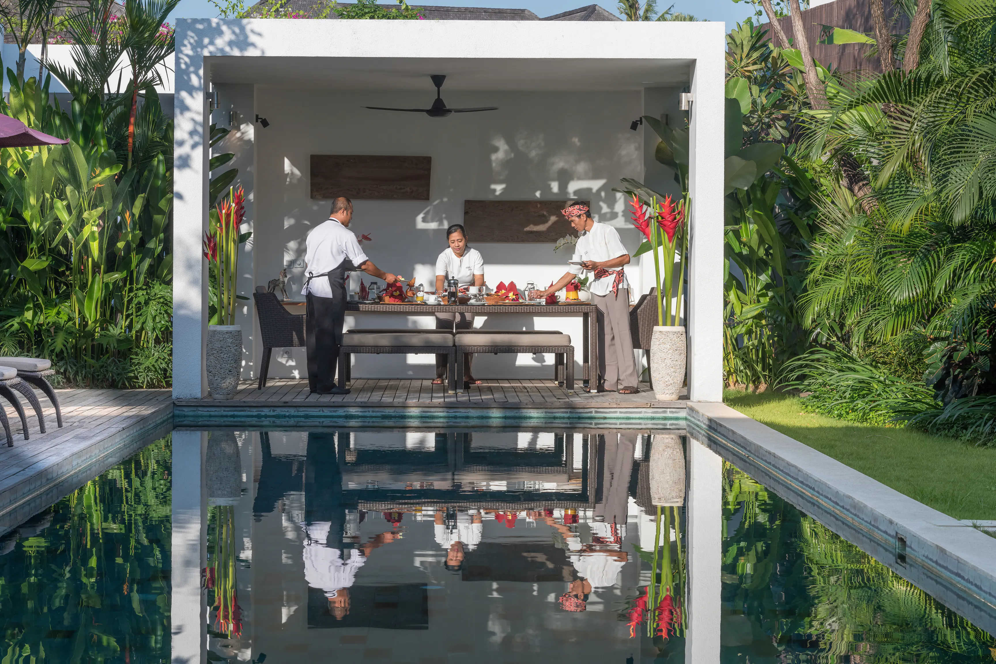 Three of the Casa Brio team laying breakfast in the poolside balé, red heliconia in stone urns either side and the whole scene mirrored the length of the pool